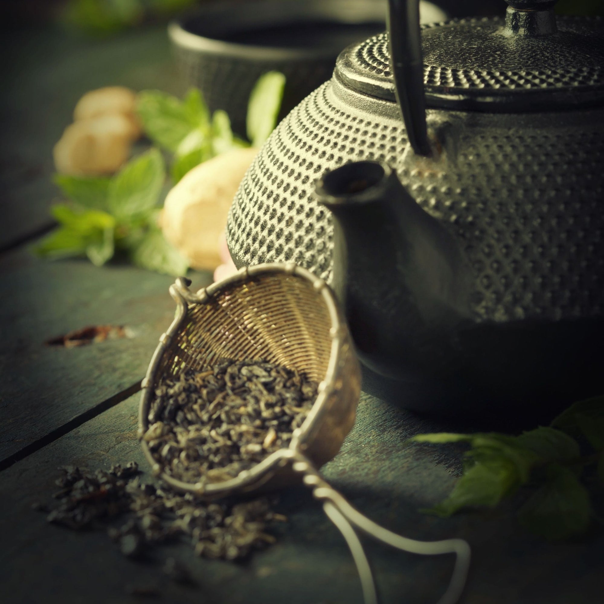 A close-up of a textured black teapot beside a tea strainer filled with loose Gamma-Aminobutyric Acid (GABA) Oolong Tea by Magic Hour. In the background, blurred elements include mint leaves and slices of ginger on a rustic wooden surface. The overall lighting is soft and warm, creating a cozy atmosphere perfect for calm nourishment.