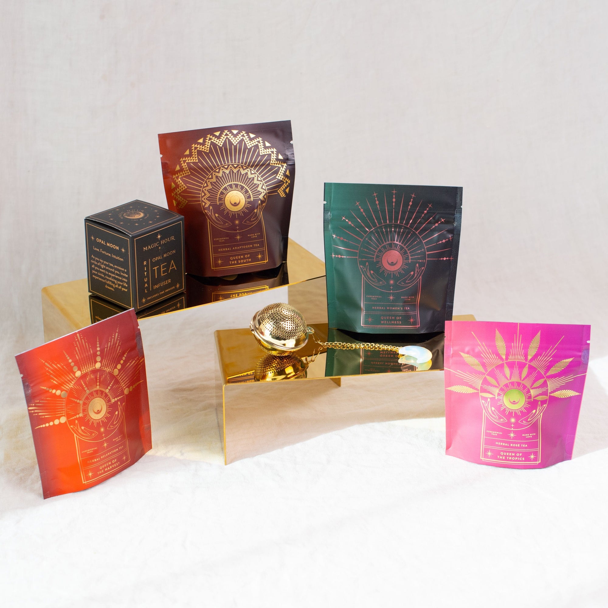 A group of four colorful tea packaging pouches with intricate designs displayed on gold platforms. There is also a gold tea infuser and a black Magic Hour box on the left side. The background is a plain, light-colored fabric, highlighting the elegance of The Queen's Sampler Set by Magic Hour.