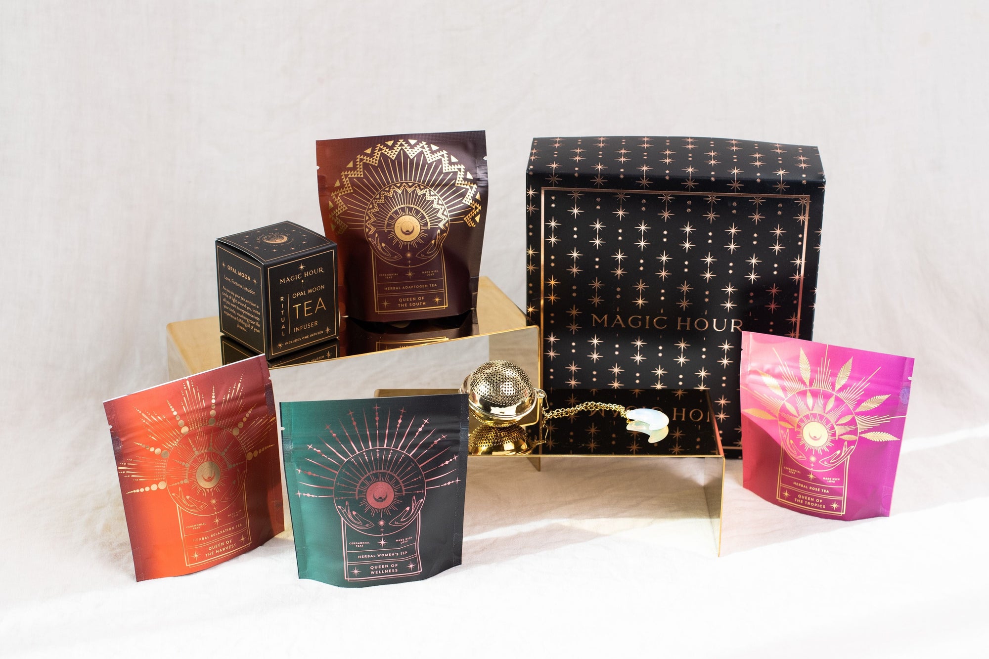 An arrangement of Magic Hour products featuring colorful packages with starry designs and a large black box adorned with gold stars. A small gold tea strainer and a brass tray are also part of the display, highlighting the elegance of their organic loose leaf tea offerings, including The Queen's Sampler Set.