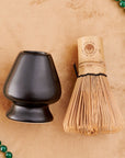 An arrangement on a beige surface features a green beaded necklace with a green tassel, a black ceramic tea bowl, and the Matte Black Chasen - Matcha Whisk Stand holding the bamboo whisk used in Japanese tea ceremonies. The necklace is arranged in a loose circle around the bowl and whisk stand.