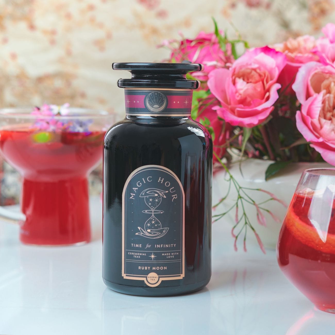 A dark glass bottle labeled "Club Magic Hour: Ruby Moon™ : Hibiscus Elderberry Tea" stands on a table. Next to it are two glasses containing red liquids with garnishes. In the background, a vase filled with vibrant, pink flowers adds a pop of color to the scene.
