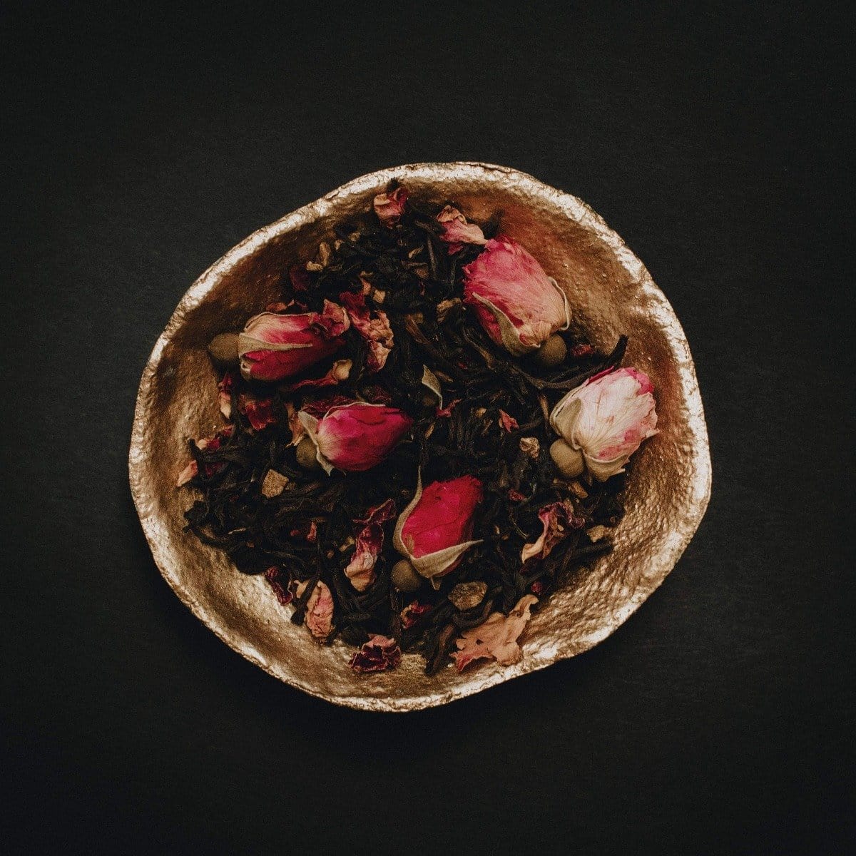 A golden bowl filled with a blend of Soulmate: Chocolate-Raspberry-Rose Black Tea for Finding &amp; Celebrating Love by Magic Hour, set against a dark background. The delicate pink and white rose buds add a touch of color to the deep brown tones of the organic tea leaves, creating an enchanting scene worthy of a magic hour.