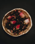 A golden bowl filled with a blend of Soulmate: Chocolate-Raspberry-Rose Black Tea for Finding & Celebrating Love by Magic Hour, set against a dark background. The delicate pink and white rose buds add a touch of color to the deep brown tones of the organic tea leaves, creating an enchanting scene worthy of a magic hour.