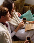 A couple sits closely together on a brown couch with teal cushions, each holding a mug of Magic Hour's Soulmate: Chocolate-Raspberry-Rose Black Tea for Finding & Celebrating Love. They are looking at an open book on the woman's lap. The woman has long dark hair and is wearing a white outfit with a beige shawl. The man has short reddish hair and is wearing a white shirt.