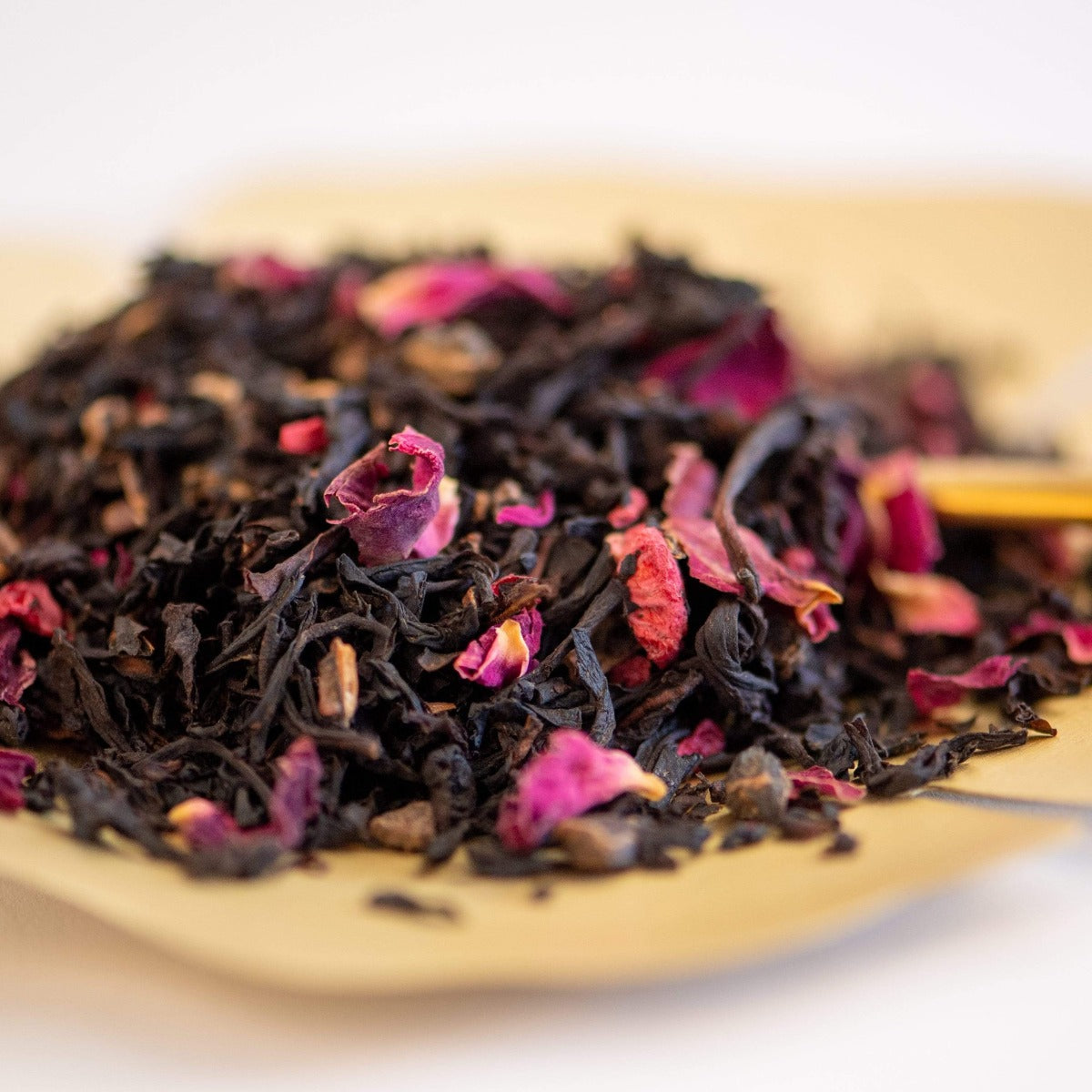 A close-up of the Enchanting Chocolate-Raspberry-Rose Tea from the Soulmate Box, showcasing loose leaf black tea blended with vibrant dried rose petals on a beige surface—ideal for a love manifestation ritual. This mixture highlights deep black leaves interspersed with bright pink and red petals, providing a delightful array of textures and colors.