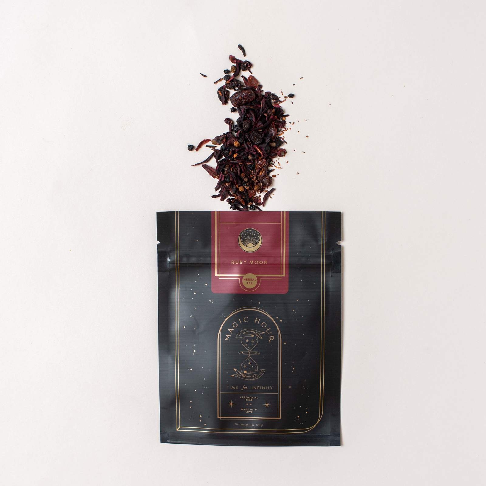 A black and gold package labeled "Magic Hour" with a red section labeled "Moon Lover: Herbal Immunitea Ritual Gift" is open at the top, spilling its organic loose leaf tea onto a white background. The dark tea leaves display a rich mix of red and brown hues.