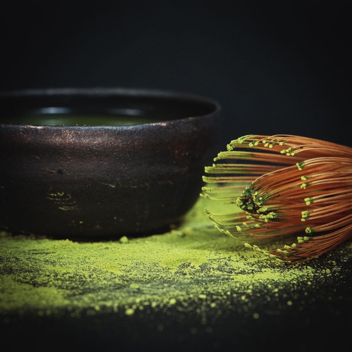 A traditional bamboo whisk sits beside a dark ceramic bowl filled with matcha tea. The Artisanal Magic Hour Matcha Whisk by Magic Hour is nestled within, while matcha powder is sprinkled around them on a dark surface, highlighting the vibrant green hue of this organic tea.