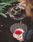 A person holding a teacup filled with red liquid in one hand and a pendulum in the other, over a decorative object on the ground. A black teapot of organic tea on a silver tray and green foliage are visible in the background. The product being showcased is the Crystal Grid: Flower of Life Tea Ceremony Altar by Alibaba.